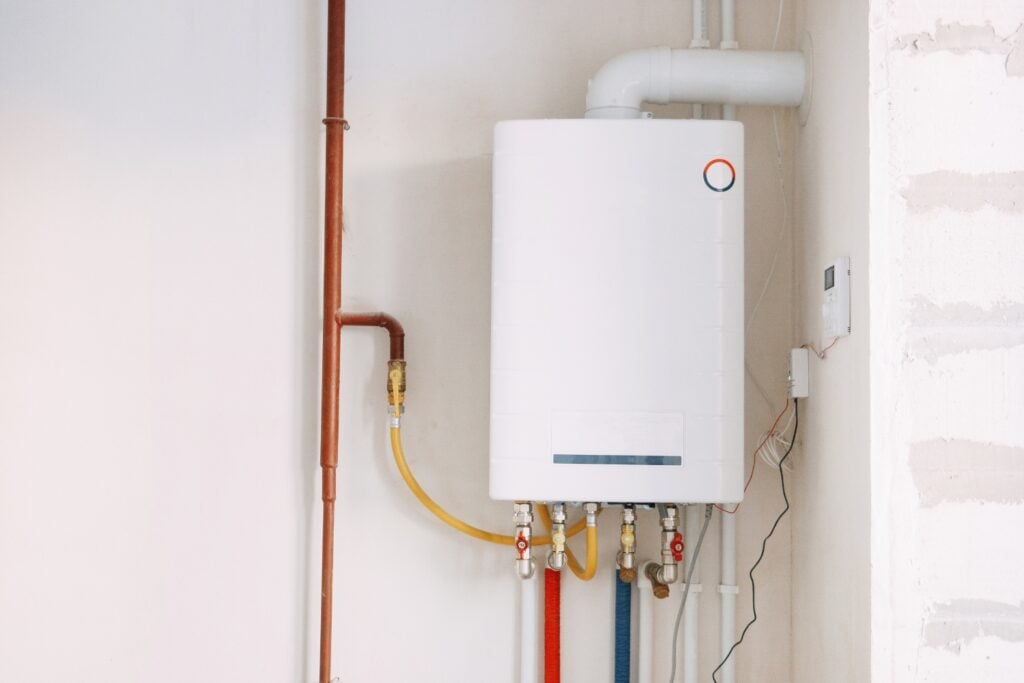 A white tankless water heater or boiler mounted on a wall with various coloured pipes and valves connected at the bottom.