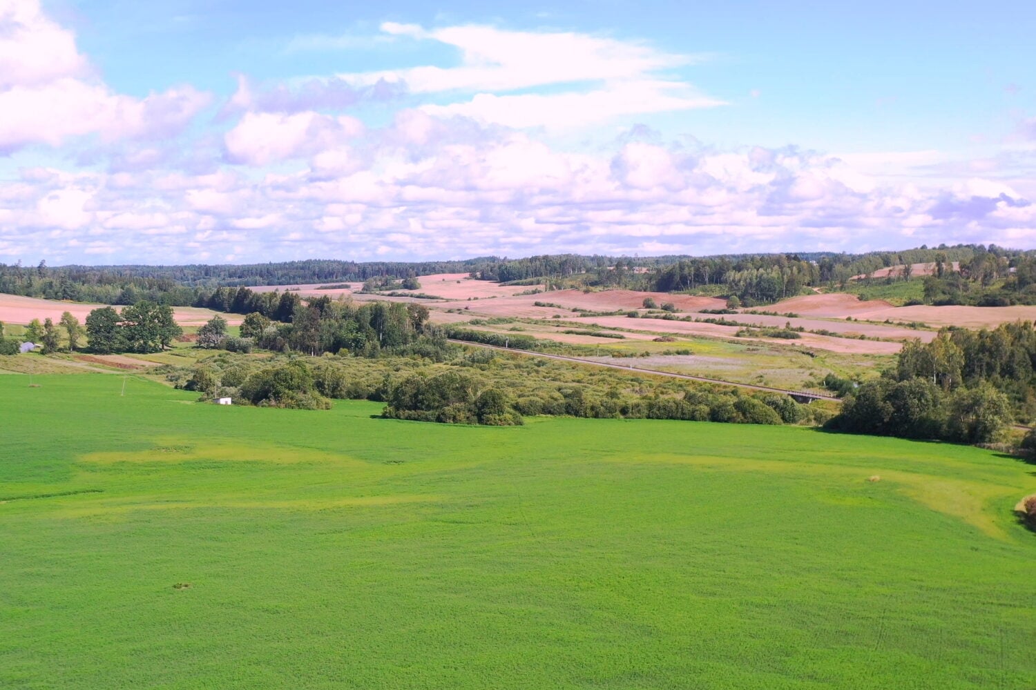 An expansive aerial view of a green rural landscape with rolling hills, patches of trees, agricultural fields, and a cloudy sky.