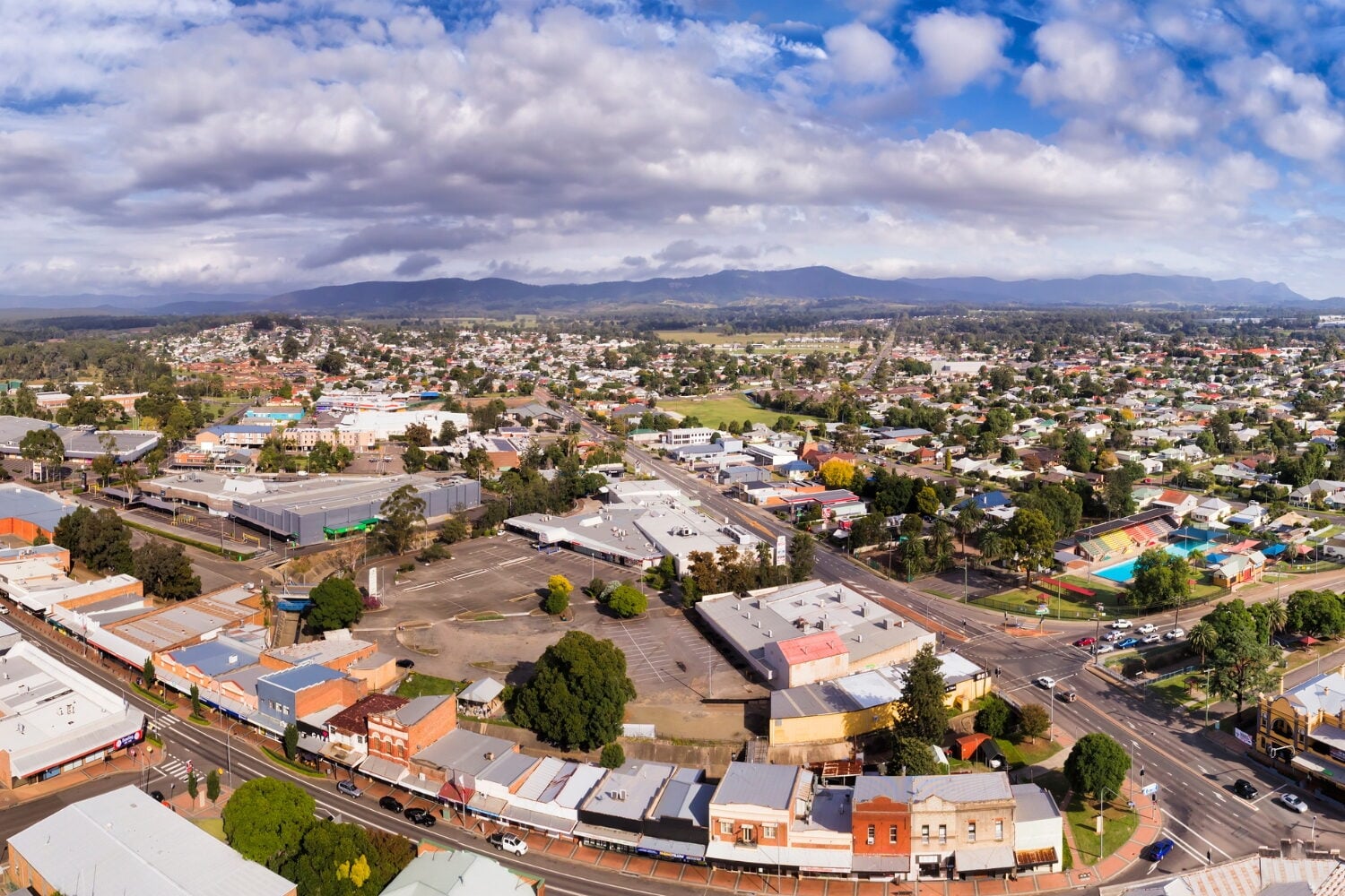A regional town centre featuring commercial buildings, streets, and residential areas stretching toward distant mountains under a partly cloudy sky.