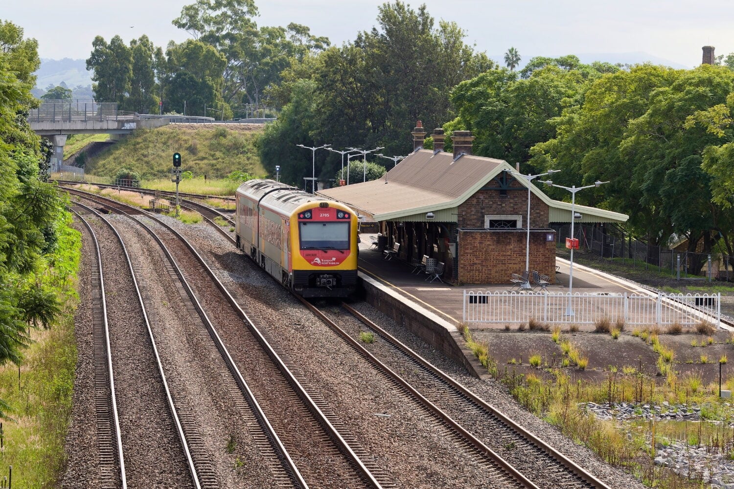 A passenger train pulling into a small brick railway station platform next to multiple tracks, surrounded by green trees.
