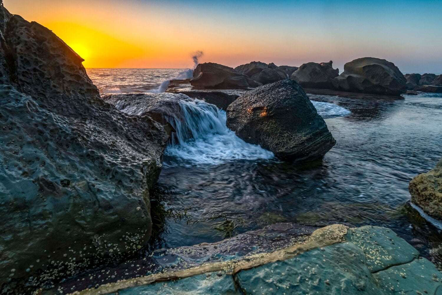 A scenic sunrise over a rocky coastline with ocean water cascading between large boulders.