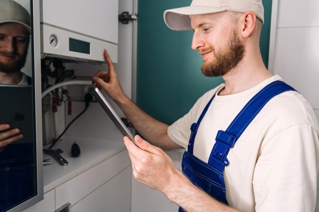 A technician with a beard, wearing a cap and blue overalls, holds a digital tablet while pressing a button on a wall-mounted boiler panel.