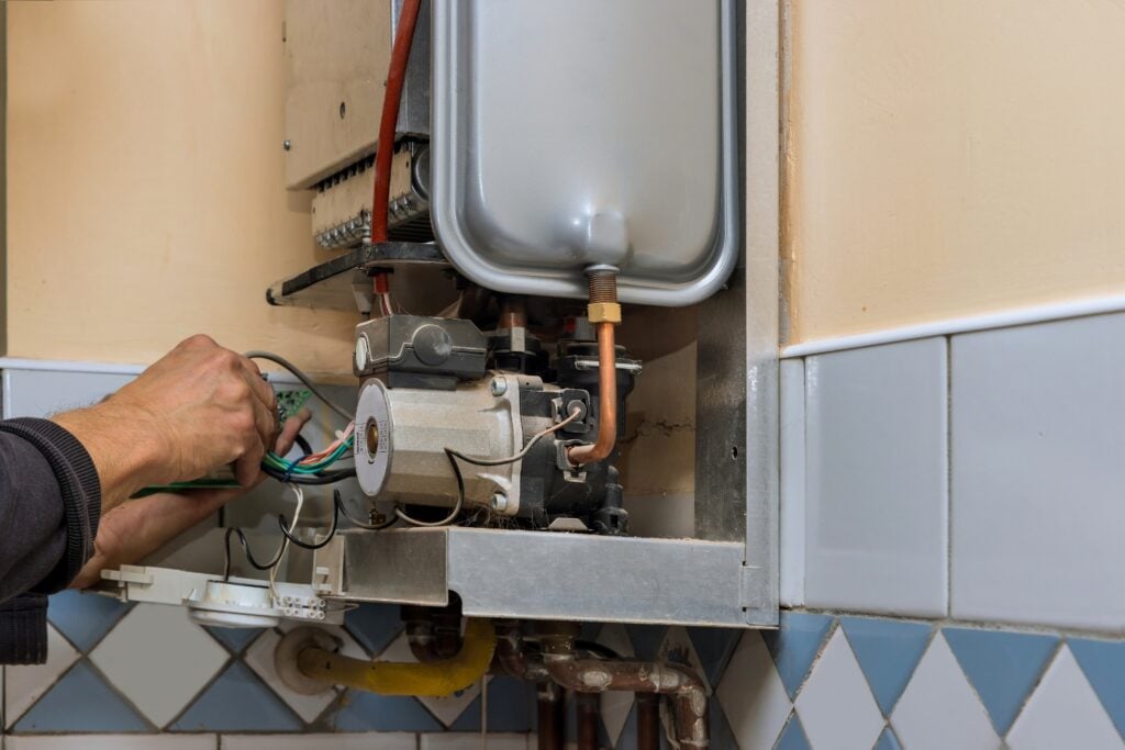 A close-up view of hands working on the internal wiring and electrical components of an open wall-mounted gas boiler system during repair or servicing.