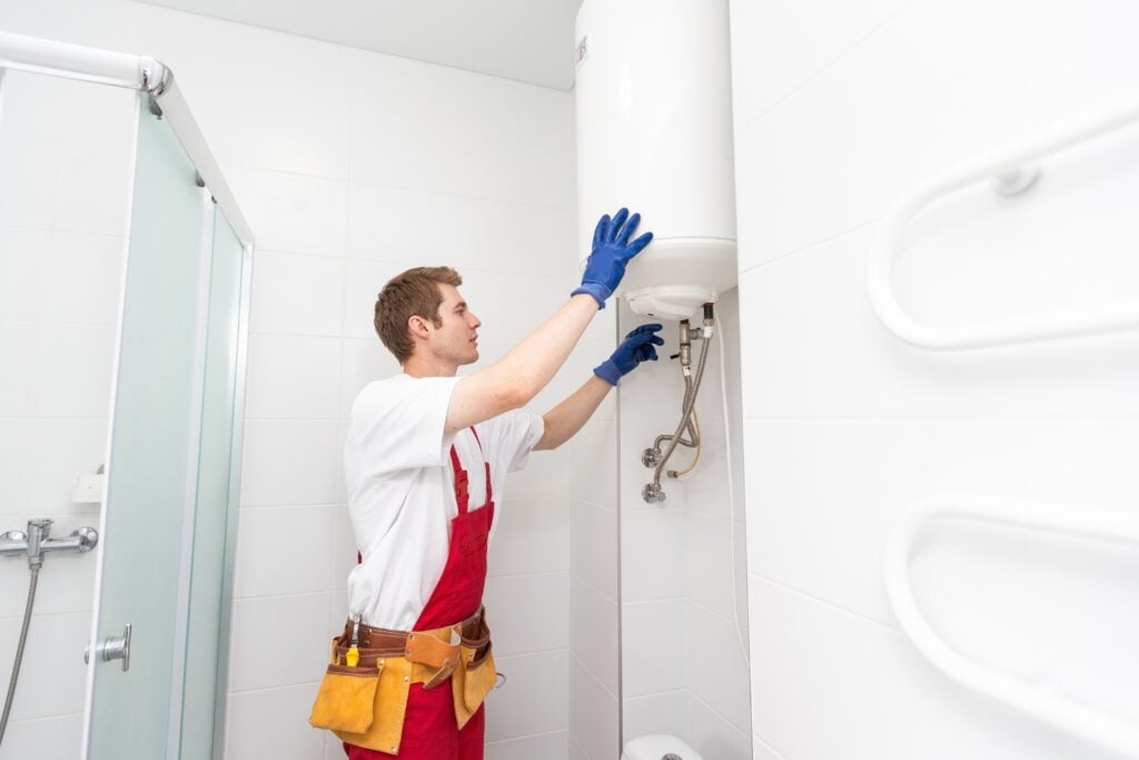 A plumber wearing a white t-shirt, red overalls, a tool belt, and blue gloves works on a wall-mounted cylindrical water heater inside a bright white bathroom.