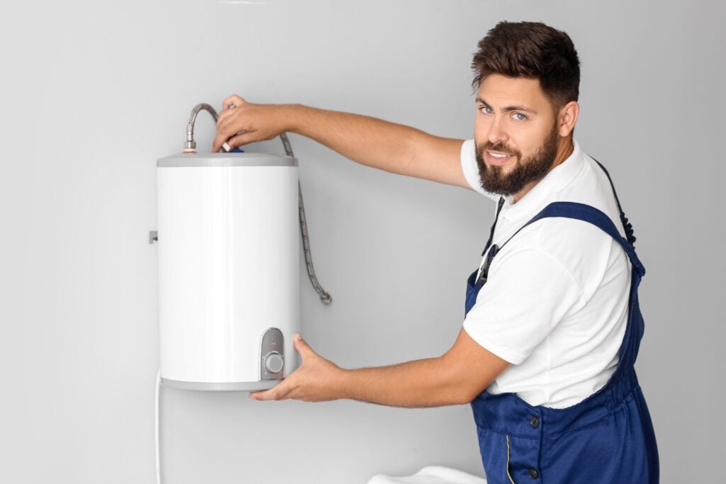 A smiling technician with a beard, wearing blue overalls, adjusts a small white cylindrical water heater mounted on a grey wall.