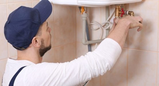 A technician wearing a blue cap and white long-sleeved shirt working on the pipe fittings beneath a white wall-mounted hot water heater unit.