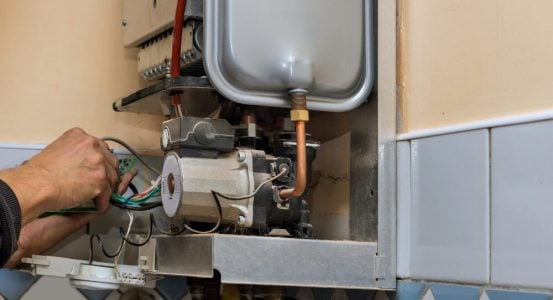 A technician's hands repairing the internal electrical wiring and circuit board panel of an opened wall-mounted gas boiler system.