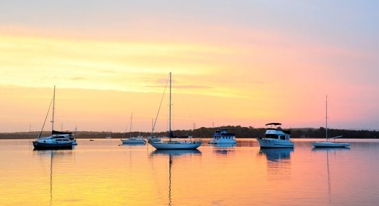 Several sailboats and motorboats anchored on a glassy, still lake during a pastel sunset.