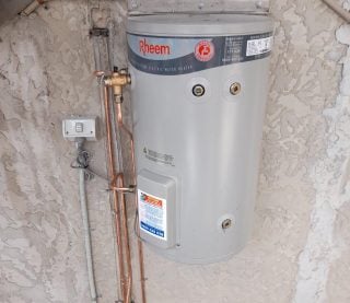 A wall-mounted grey Rheem electric water heater on a textured exterior wall with copper plumbing.