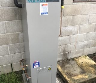 A tall, rectangular Vulcan Freeloader gas hot water system installed outside against a concrete block wall.