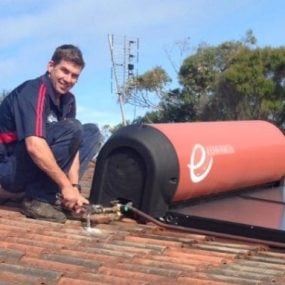 A technician in a dark blue polo with red stripes smiling while using a wrench to work on the plumbing