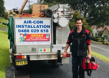 A technician in a black and red jacket standing next to the rear of a Hogan Hot Water & Air Conditioning service truck