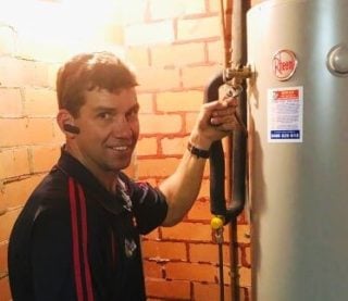Home » Hot water system A technician in a dark blue polo shirt smiling while adjusting a valve on a tall, silver Rheem hot water heater