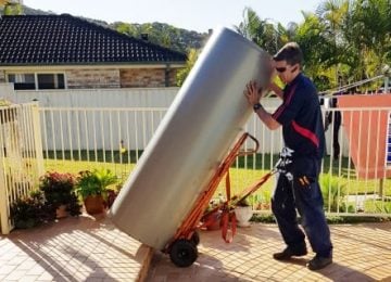 A technician in a dark blue uniform uses an orange hand truck to carefully move a large, silver hot water tank