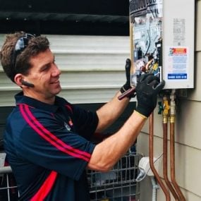 A technician in a dark blue polo shirt with red stripes wearing black gloves while inspecting the internal components of a wall-mounted continuous flow hot water heater.