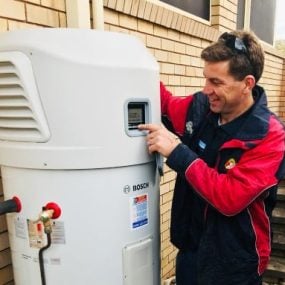A technician in a red and dark blue jacket smiling while operating the digital control panel of a large, white Bosch heat pump hot water system