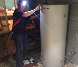 A smiling technician wearing a headlamp and earmuffs stands next to a tall, cream-coloured Thermann hot water storage tank
