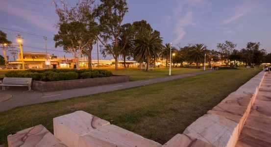 A tranquil park scene at dusk featuring a paved walkway lined with palm trees and streetlights